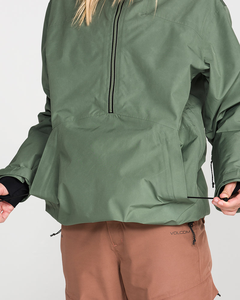 A person wearing a green Volcom Women’s Rowan Insulated Gore Pullover and brown Volcom shorts tightens the drawstring at the pullover’s hem, with their face not visible.