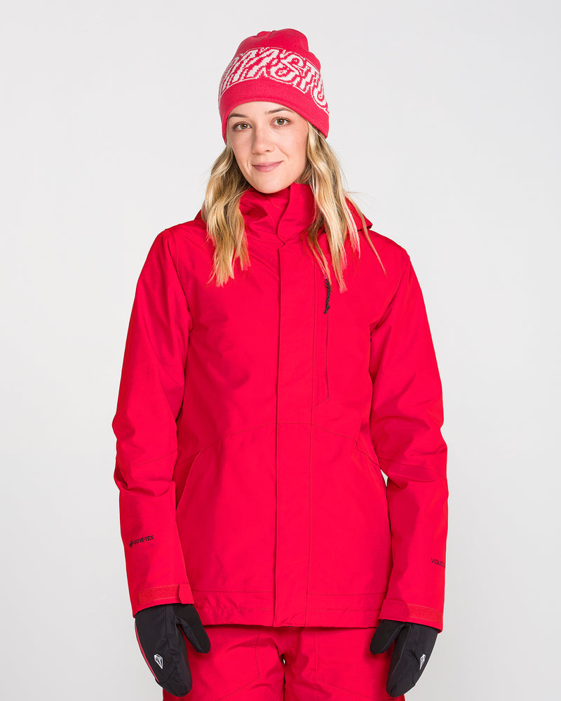 A person is wearing the Volcom Women’s Eras Gore Insulated Jacket in red, paired with red pants, black gloves, and a red knit hat with a white pattern, standing against a plain white background.