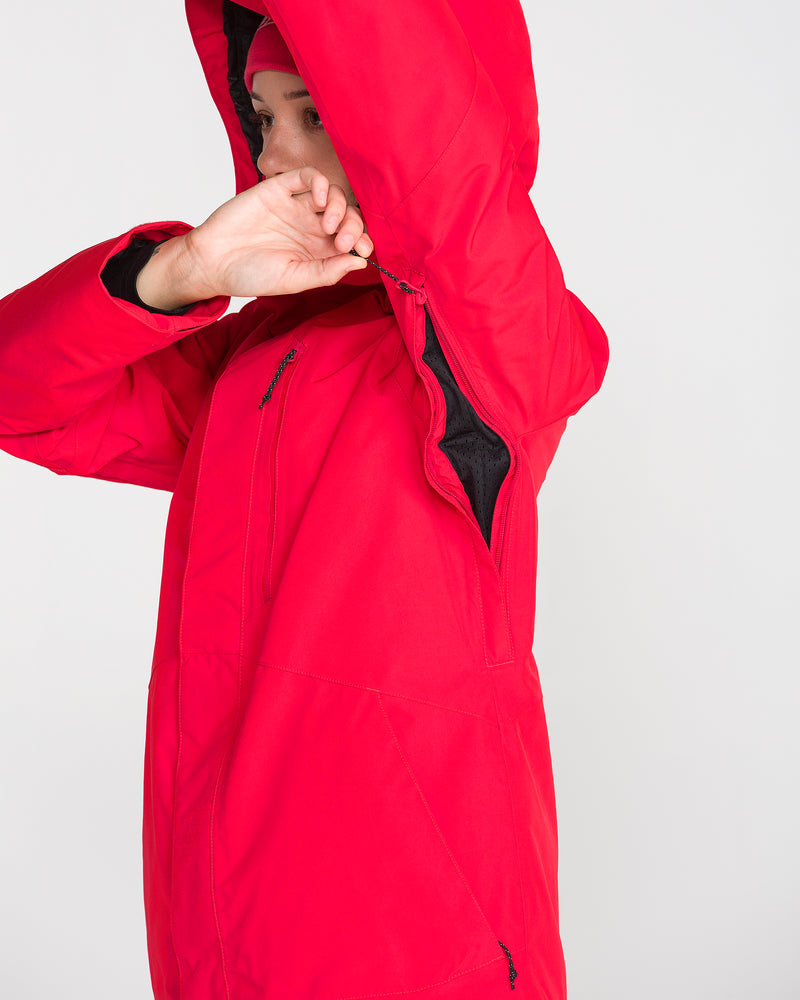A person wearing the Volcom Women’s Eras Gore Insulated Jacket in bright red lifts their arm to reveal a zippered underarm vent. Their face is partially hidden by the jacket’s hood and their raised arm, set against a plain, light background.
