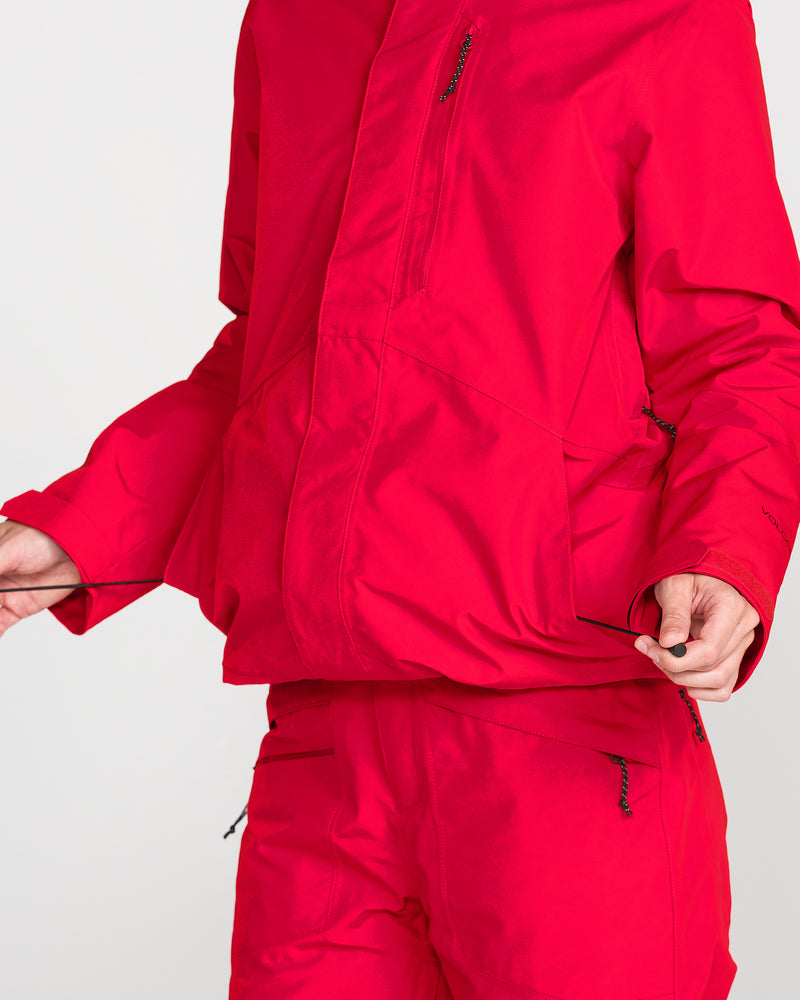 A person is shown wearing the Volcom Women’s Eras Gore Insulated Jacket in bright red, pulling at the front zipper. Only the torso and part of the arms are visible against a plain white background.