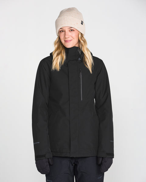 A person wearing a beige knit beanie, gloves, and the Volcom Women’s Eras Gore Insulated Jacket stands facing forward against a plain white background, smiling gently.