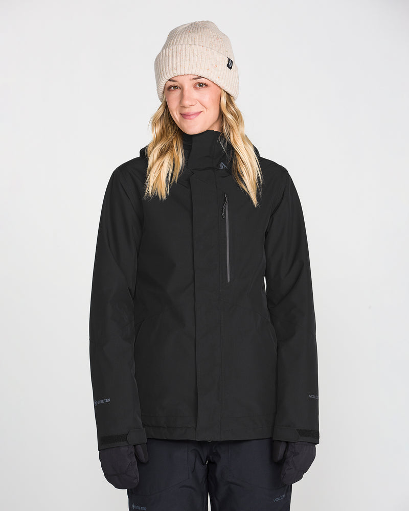 A person wearing a beige knit beanie, gloves, and the Volcom Women’s Eras Gore Insulated Jacket stands facing forward against a plain white background, smiling gently.
