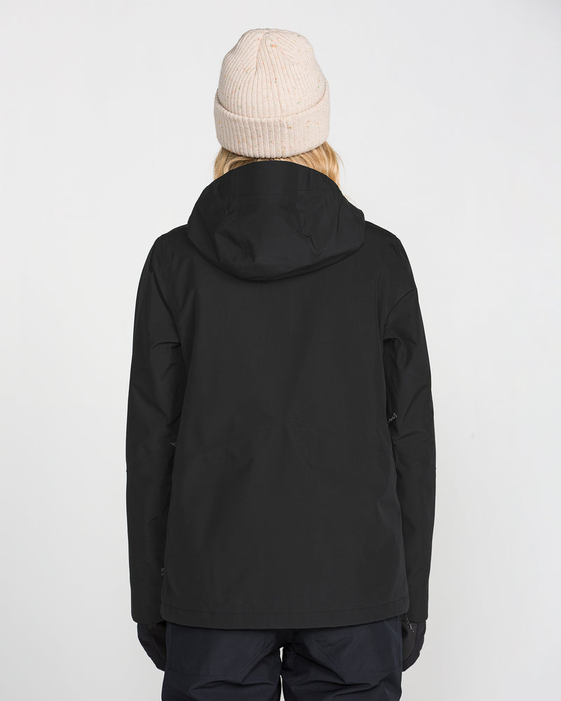 A person is standing with their back to the camera against a plain light background, wearing the Volcom Women’s Eras GORE Insulated Jacket in black, paired with black pants and a light beige knit beanie.