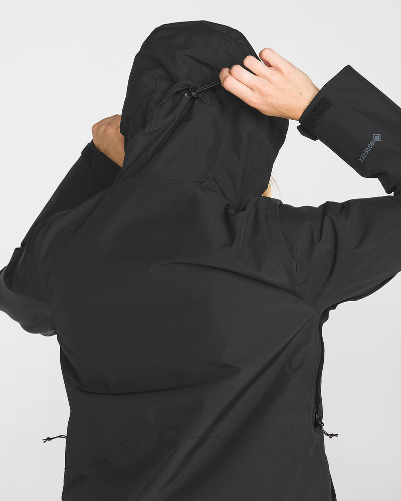 A person is seen from the back, pulling up the hood of the Volcom Women’s Eras Gore Insulated Jacket in black, with only their arm, hand, and part of their head visible against a plain light background.