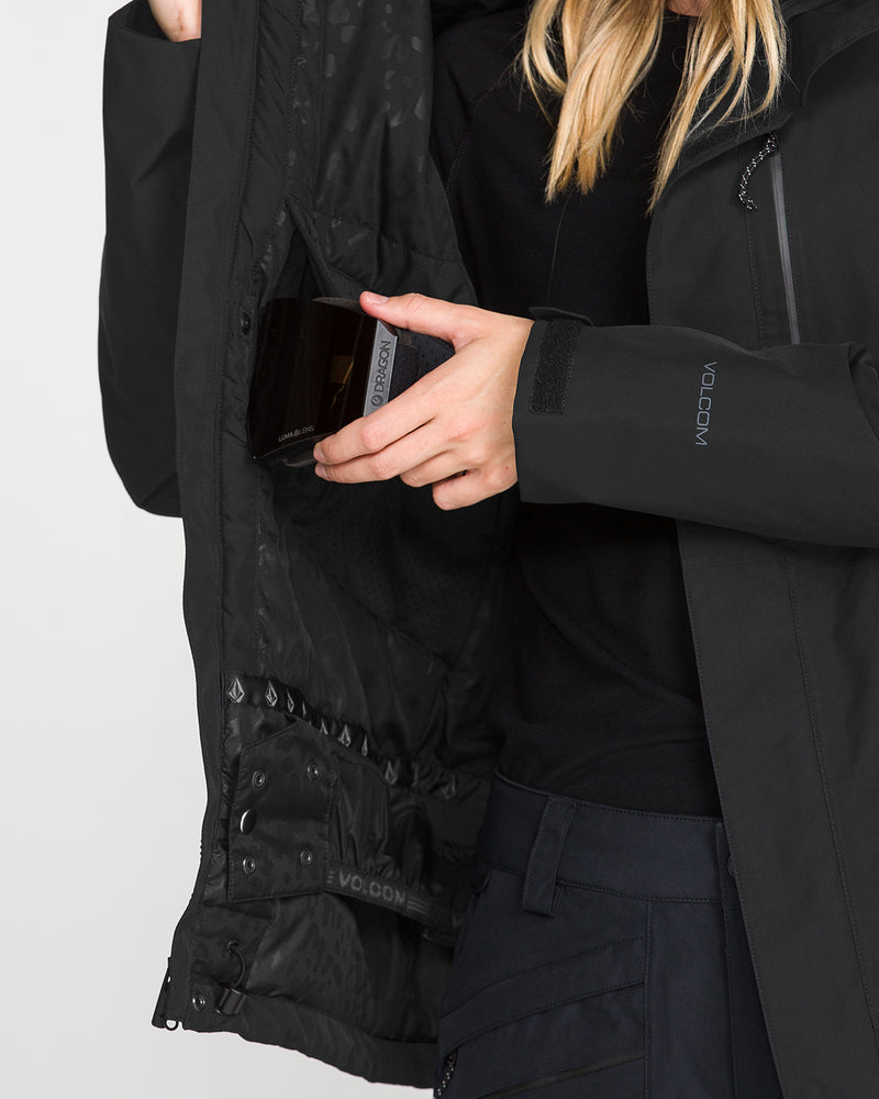 A person wearing the Volcom Women’s Eras Gore Insulated Jacket in black places a black rectangular object into an inner pocket of the jacket, with only their hands and part of their blonde hair visible.