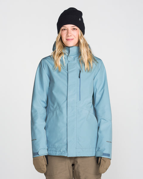 A woman wearing the Volcom Women’s Eras Gore Insulated Jacket in light blue, paired with a black knit beanie and brown gloves, stands facing forward against a plain white background, smiling slightly.