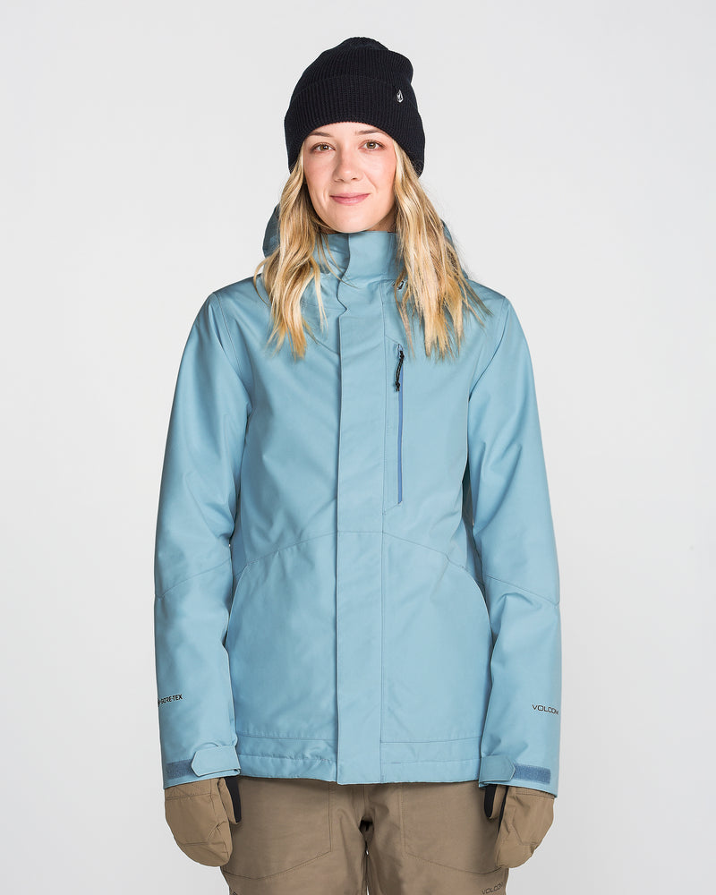 A woman wearing the Volcom Women’s Eras Gore Insulated Jacket in light blue, paired with a black knit beanie and brown gloves, stands facing forward against a plain white background, smiling slightly.