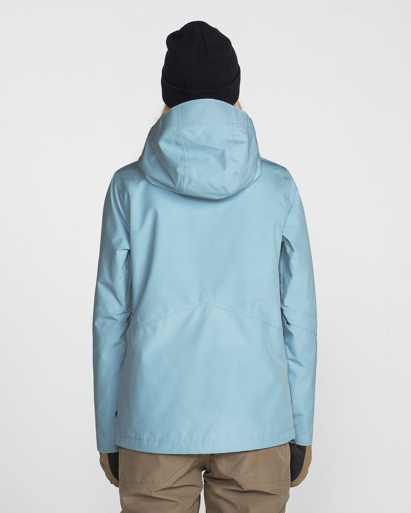 A person is seen from behind, standing against a plain white background, wearing the Volcom Women’s Eras Gore Insulated Jacket in light blue, along with a black beanie, brown pants, and black gloves.