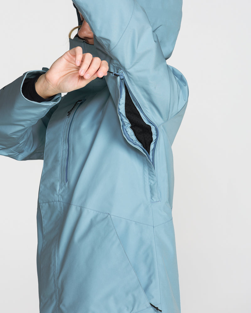 A woman wearing the Volcom Women’s Eras Gore Insulated Jacket in light blue unzips the underarm ventilation opening. The side view highlights the jacket’s unzipped vent feature.