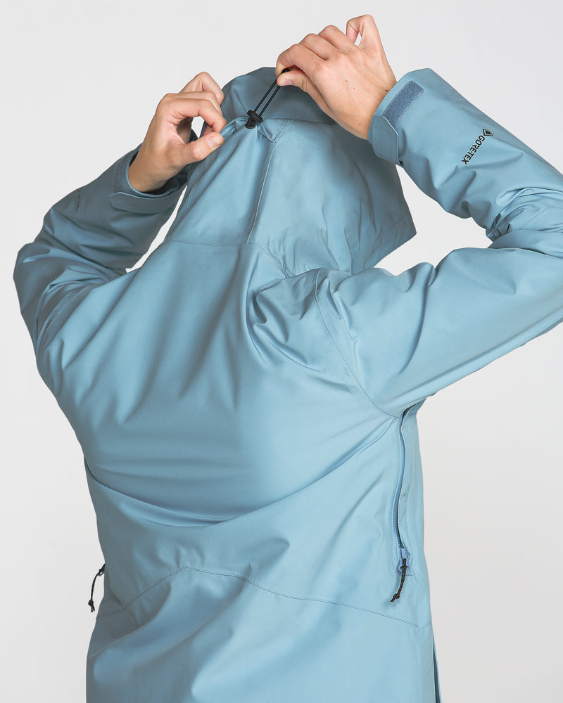 A woman wearing the Volcom Women’s Eras Gore Insulated Jacket in blue adjusts the hood using the back drawcord. The photo, captured from behind, showcases the jacket’s functional features and her hand movements, emphasizing the premium quality of this waterproof outerwear.