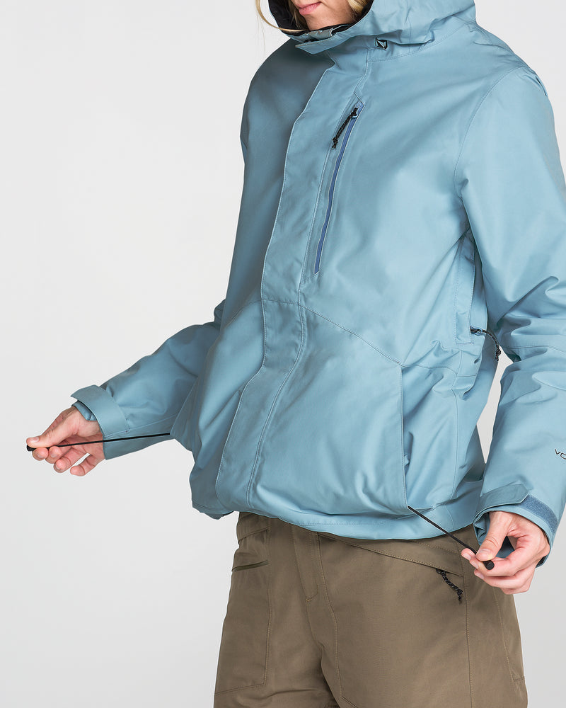 A person wearing the Volcom Women’s Eras Gore Insulated Jacket in light blue, featuring a zippered chest pocket, paired with brown pants and standing against a plain white background. The individual’s face is partially out of frame.