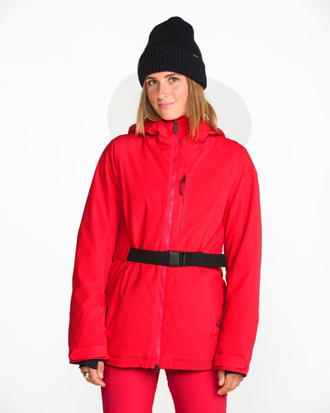 A person stands facing forward against a plain white background, wearing a bright red Volcom Women’s Alpine All Day Insulated Jacket made with 10K/10K V-Science fabric and 4-way stretch, paired with a black belt, matching red pants, and a black knit beanie.