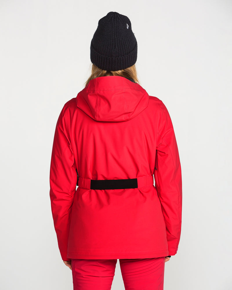 A person facing away, wearing a bright red Volcom Women’s Alpine All Day Insulated Jacket, red 4-way stretch pants, and a black knit beanie, stands against a plain light-colored background.