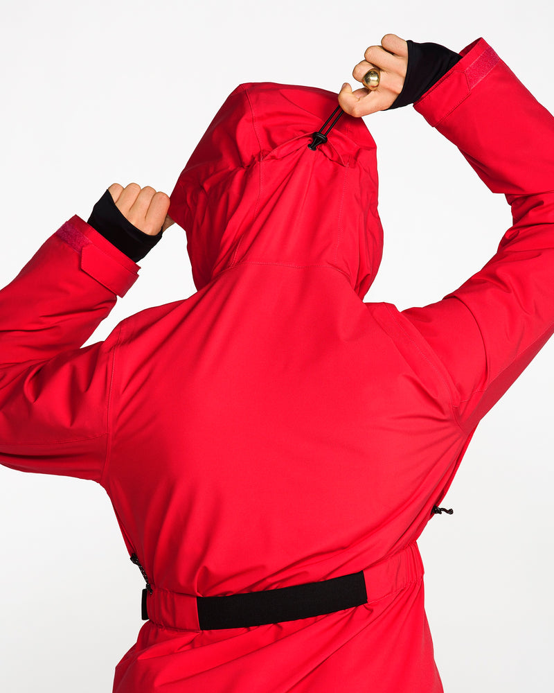 A person is seen from behind adjusting the hood of the vibrant red Volcom Women’s Alpine All Day Insulated Jacket, which features 4-way stretch and 10K/10K V-Science technology. The jacket stands out against a plain white background.