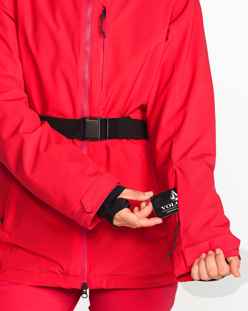 A person wearing a vibrant red Volcom Women’s Alpine All Day Insulated Jacket with a black belt reveals an inner sleeve tag featuring the Volcom logo. The jacket’s 4-way stretch fabric and 10K/10K V-Science technology deliver comfort and protection, while the wearer’s thumb passes through a sleeve thumbhole. The background is plain white.