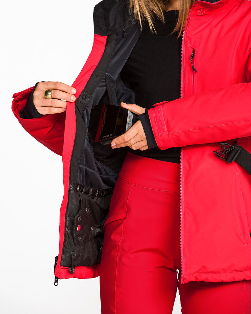 A person wearing a red Volcom Women’s Alpine All Day Insulated Jacket and matching pants opens an inner pocket to reveal a smartphone stored inside. Their head is out of frame against a plain white background. The jacket’s 10K/10K V-Science fabric and 4-way stretch construction enhance the versatility and subtle detail of this look.