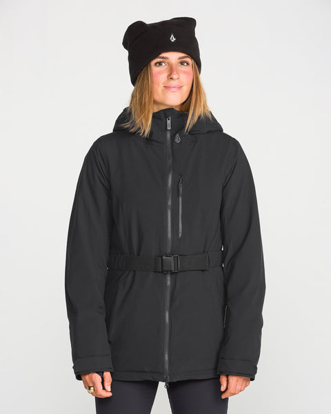 A woman wearing the Volcom Women’s Alpine All Day Insulated Jacket with a belt and a black beanie stands facing forward against a plain white background, smiling slightly.