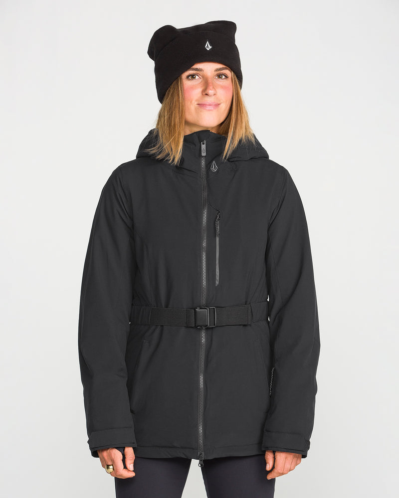 A woman wearing the Volcom Women’s Alpine All Day Insulated Jacket with a belt and a black beanie stands facing forward against a plain white background, smiling slightly.