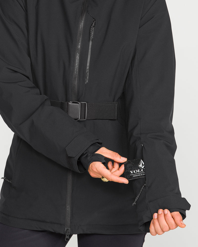 A person models the Volcom Women’s Alpine All Day Insulated Jacket in black, displaying the inside sleeve pocket with a visible Volcom logo tag. The jacket features a belt and a zippered chest pocket, with only the torso and arms shown.