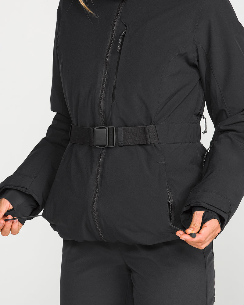 A person wearing the Volcom Women’s Alpine All Day Insulated Jacket in black and matching black pants pulls on the jacket’s drawstring; the image highlights the torso, showcasing features like the coat’s belt and zippers.
