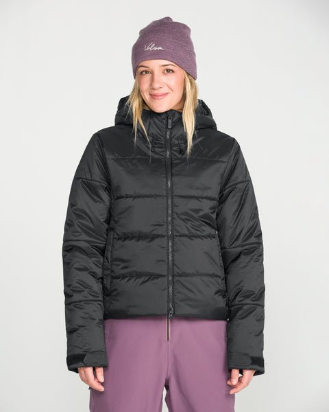 A person wearing a purple beanie, purple pants, and the Volcom Women’s Avalon Puff Jacket with recycled insulation stands facing forward against a plain white background, smiling slightly.