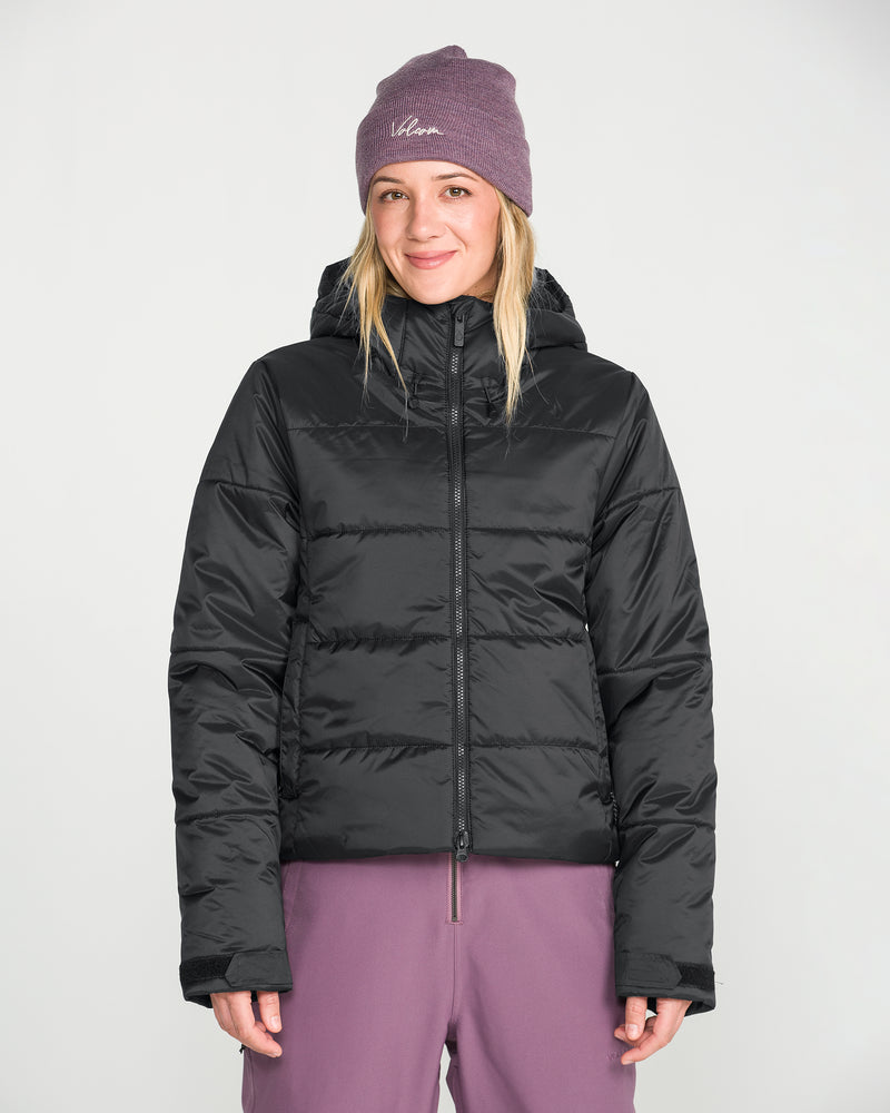 A person wearing a purple beanie, purple pants, and the Volcom Women’s Avalon Puff Jacket with recycled insulation stands facing forward against a plain white background, smiling slightly.