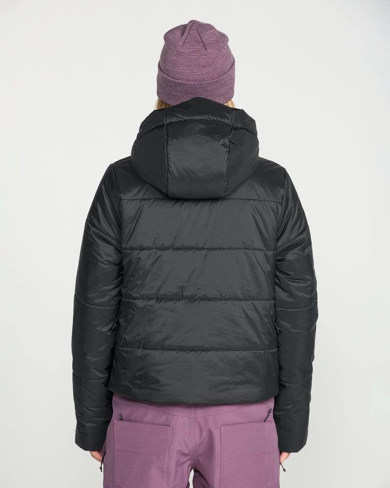 A person stands with their back to the camera, dressed in Volcom’s Women’s Avalon Puff Jacket—a black, waterproof jacket with recycled insulation—paired with mauve pants and a matching mauve beanie, set against a plain white background.