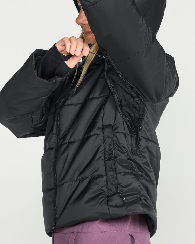 A person models the Volcom Women’s Avalon Puff Jacket—a black puffer featuring recycled insulation, a hood, and a partially unzipped side revealing its ventilation detail—styled with purple pants; their arm is raised and face partly obscured against a plain backdrop.