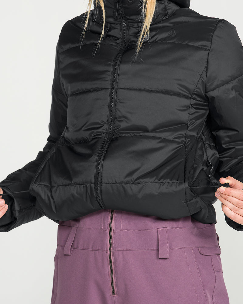 A person wearing the Volcom Women’s Avalon Puff Jacket, a black puffer crafted with recycled insulation, paired with high-waisted purple pants; shown from shoulders to hips, hands gently pulling at the sides of the jacket.