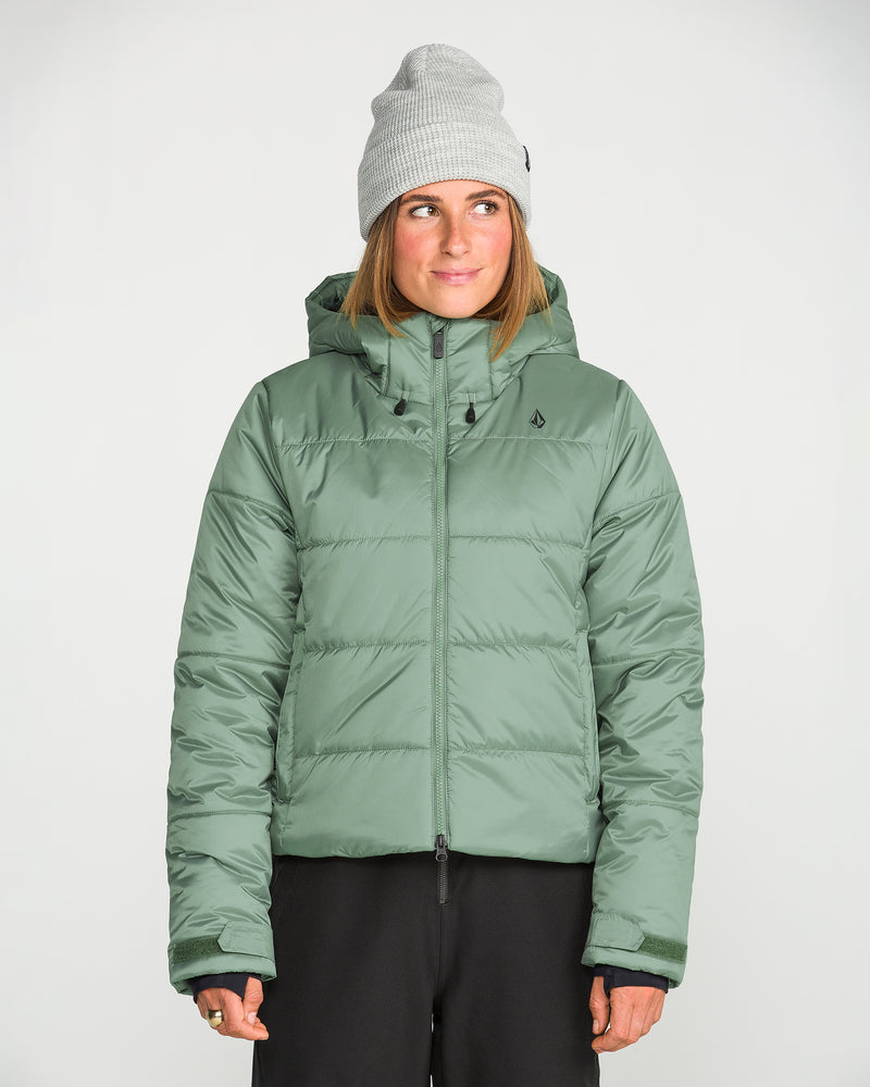A woman stands against a plain white background, looking slightly off to the side with a slight smile. She is wearing a light gray beanie, black pants, and the Volcom Women’s Avalon Puff Jacket in sage green, which features recycled insulation.