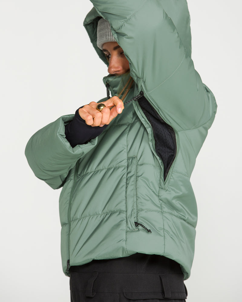 Wearing a green Volcom Women’s Avalon Puff Jacket with recycled insulation and a gray beanie, a person zips a side vent on their jacket. The photo is cropped to display their torso and arms, with the hood partially up.