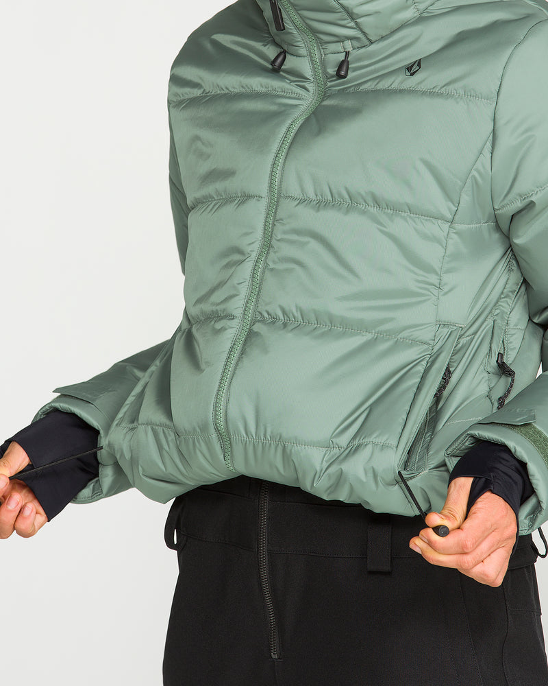 Wearing the Volcom Women’s Avalon Puff Jacket in green, with thumbholes at the sleeves, a person holds the jacket’s hem and zipper to display its pockets alongside part of black pants; this women’s jacket features recycled insulation. The head and most of the face are out of frame.