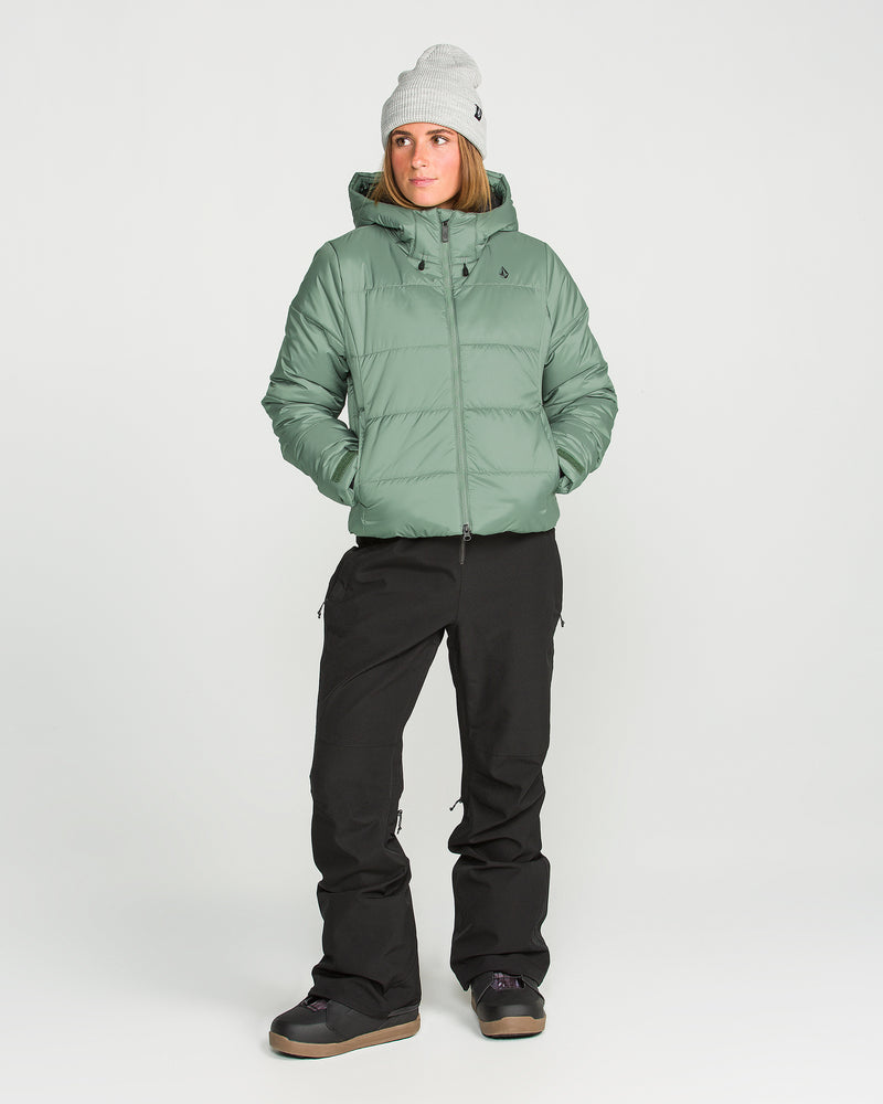 A person stands against a plain white background wearing the Volcom Women’s Avalon Puff Jacket—a stylish light green women’s jacket with recycled insulation—paired with black snow pants, a gray beanie, and black winter boots.
