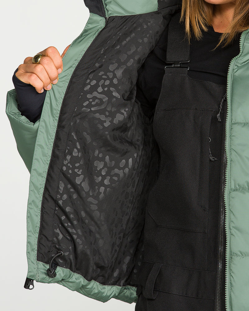 A person dressed in black overalls holds open the green Volcom Women’s Avalon Puff Jacket, showcasing its black inner lining accented with a subtle leopard print pattern. This jacket features recycled insulation for enhanced warmth and sustainability.