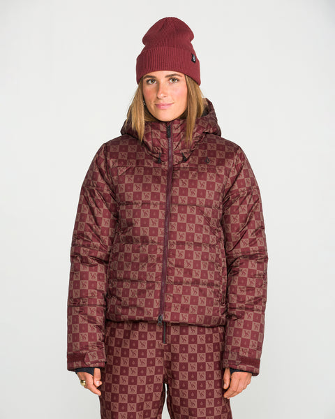 A woman stands against a plain white background wearing a matching maroon beanie, Volcom Women’s Avalon Puff Jacket with recycled insulation, and checkerboard patterned pants.