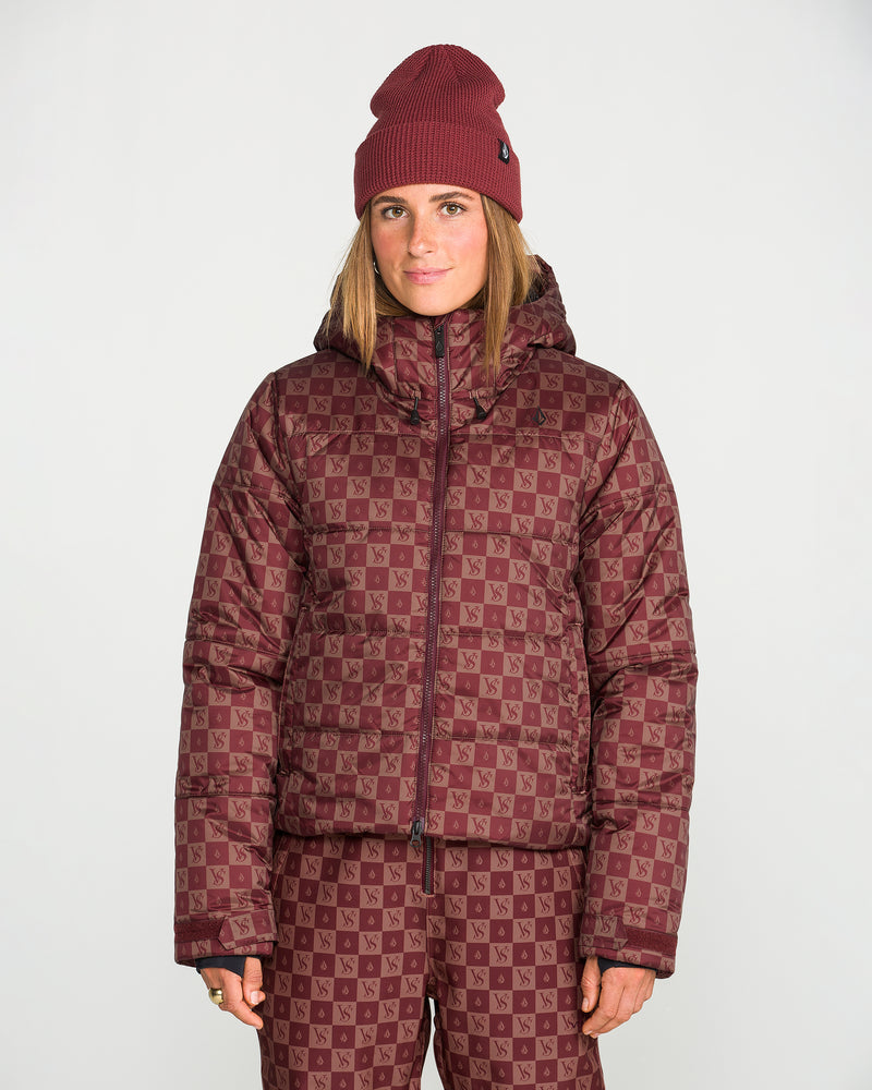 A woman stands against a plain white background wearing a matching maroon beanie, Volcom Women’s Avalon Puff Jacket with recycled insulation, and checkerboard patterned pants.