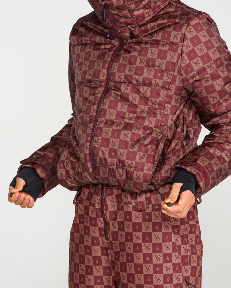A person is seen wearing a maroon matching jacket and pants set with a checkered pattern featuring VS initials, holding the drawstrings of the jacket. The women’s jacket—Volcom’s Avalon Puff Jacket—is made with recycled insulation for warmth and sustainability. The person’s face is not visible.