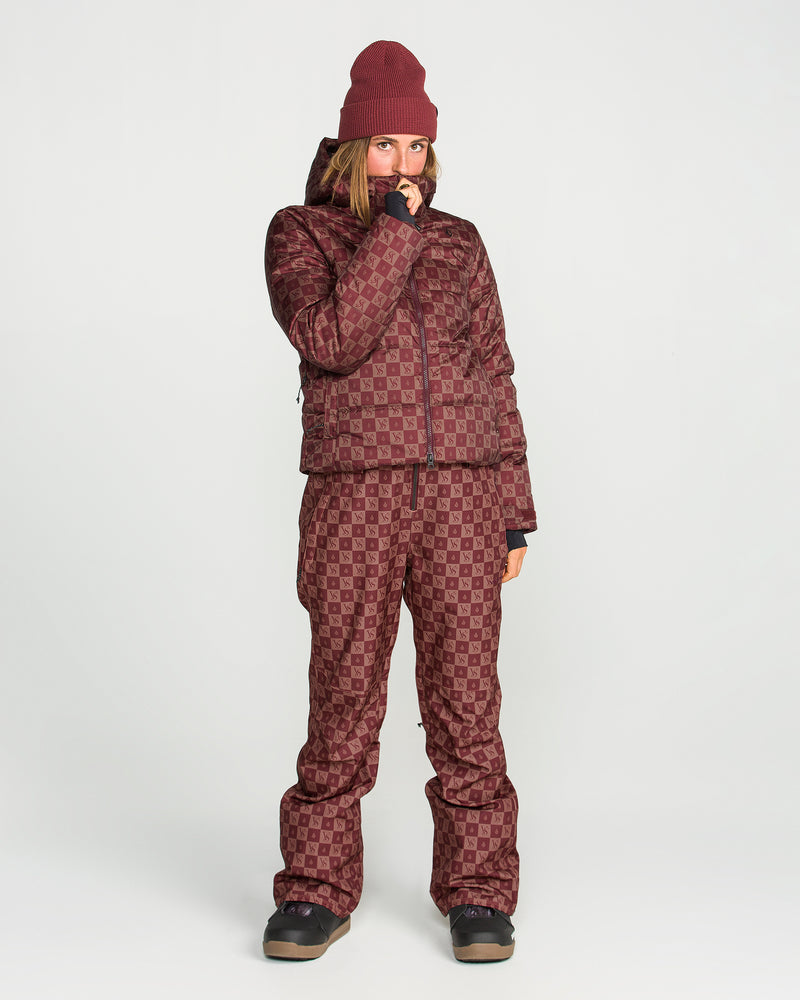 A person dressed in a maroon patterned winter ensemble stands against a plain white background, featuring the Volcom Women’s Avalon Puff Jacket with recycled insulation, matching pants and beanie, and black boots.