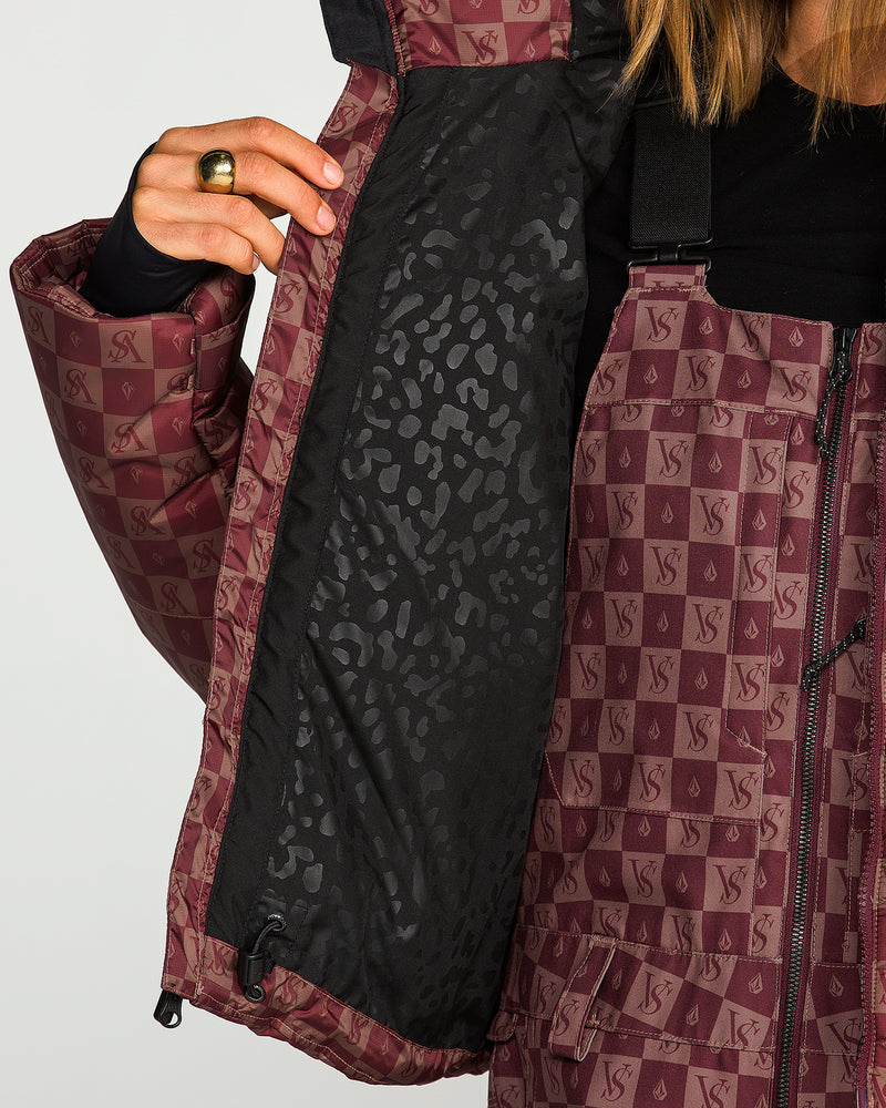 A person opens a maroon patterned Women’s Avalon Puff Jacket by Volcom, revealing its black, subtly patterned inner lining. The jacket features recycled insulation and a repeated VS logo and is worn by someone sporting a ring on their finger.