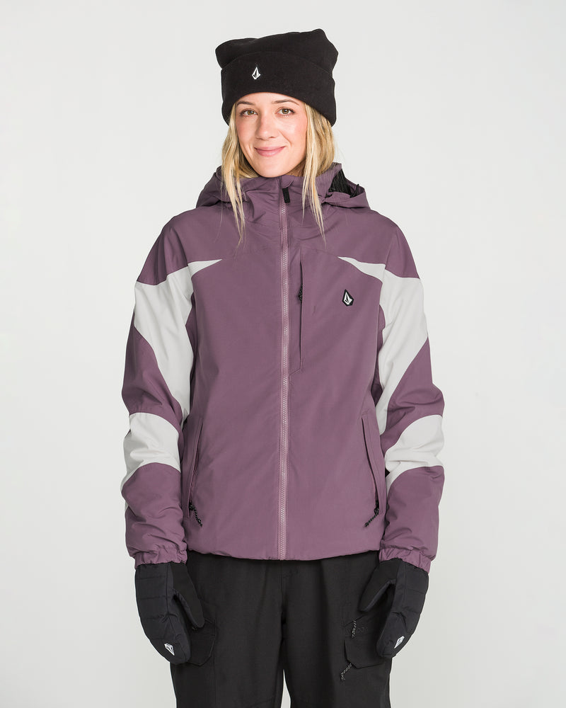 A woman is standing facing forward against a plain white background, smiling slightly, while wearing the Volcom Women’s Shelter 3D Stretch Jacket in purple and gray, paired with black gloves and a black beanie.