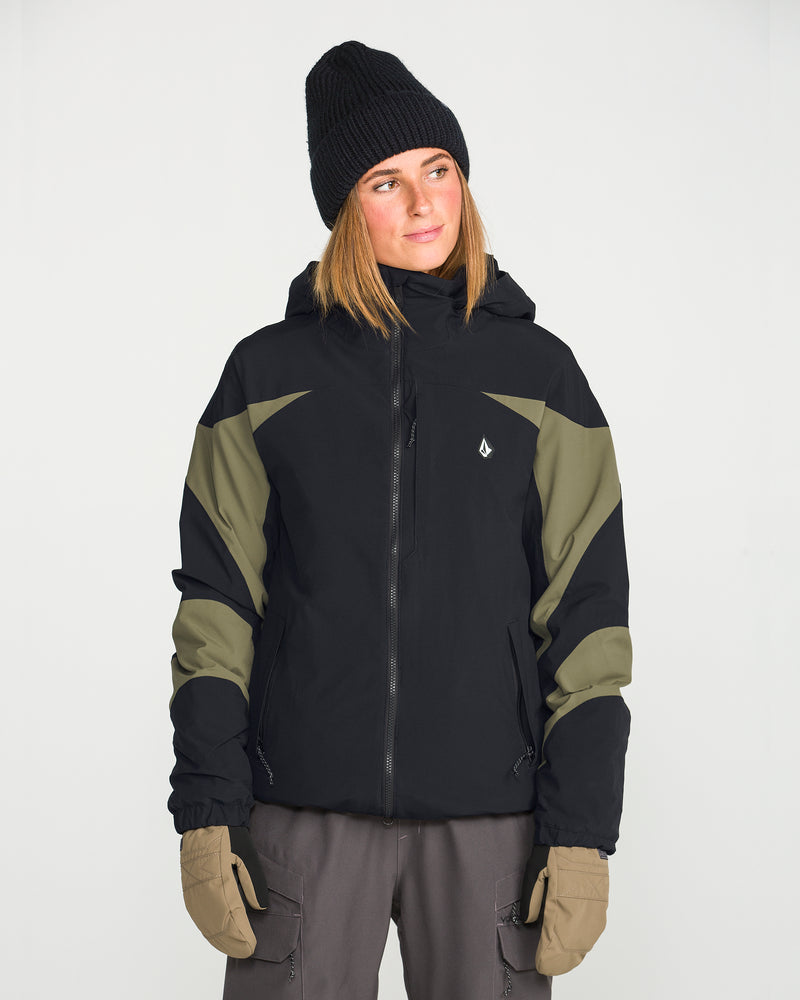 A person wearing a black beanie, tan gloves, gray pants, and the Volcom Women’s Shelter 3D Stretch Jacket in black and olive green with 20K/20K protection stands against a plain white background, looking slightly to the right.