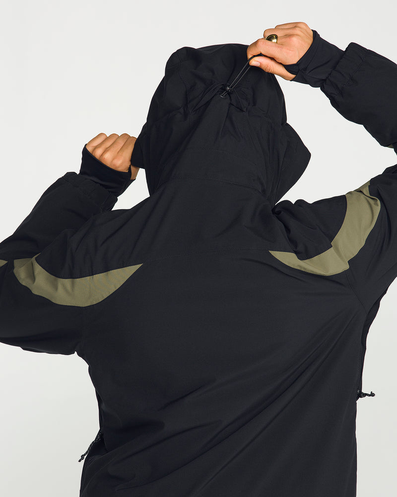 A person faces away from the camera against a plain light background, pulling up the hood of the Volcom Women’s Shelter 3D Stretch Jacket—a black jacket with olive green accents designed with 3D Stretch fabric for enhanced mobility.