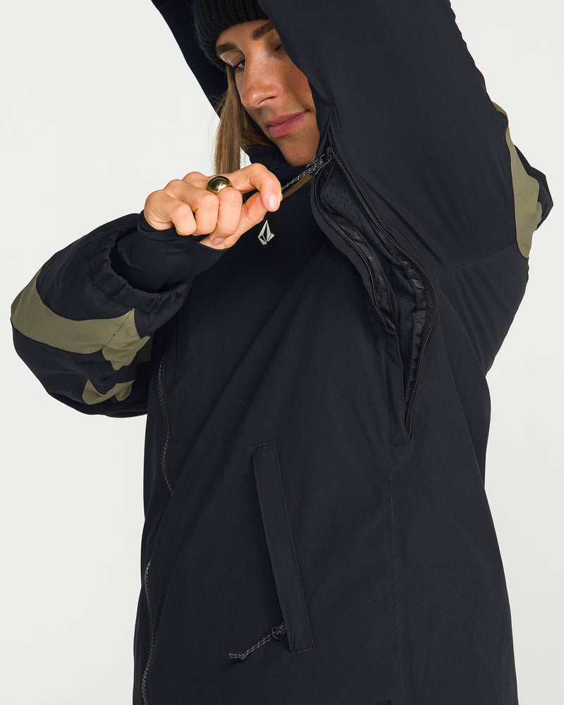 Wearing the Volcom Women’s Shelter 3D Stretch Jacket—a black piece featuring green sleeve accents—a person unzips an underarm zipper to reveal its low-loft insulated interior lining. Partially visible, their face is framed by a black beanie.