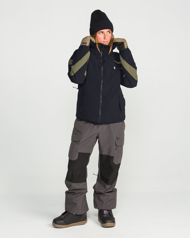 A person is shown wearing the Volcom Women’s Shelter 3D Stretch Jacket—a black winter jacket with green stripes, engineered with advanced 20K/20K defense and low-loft insulation for optimal warmth and protection. They style it with gray cargo pants featuring black knee patches, black boots, and a black beanie, standing against a plain white background.