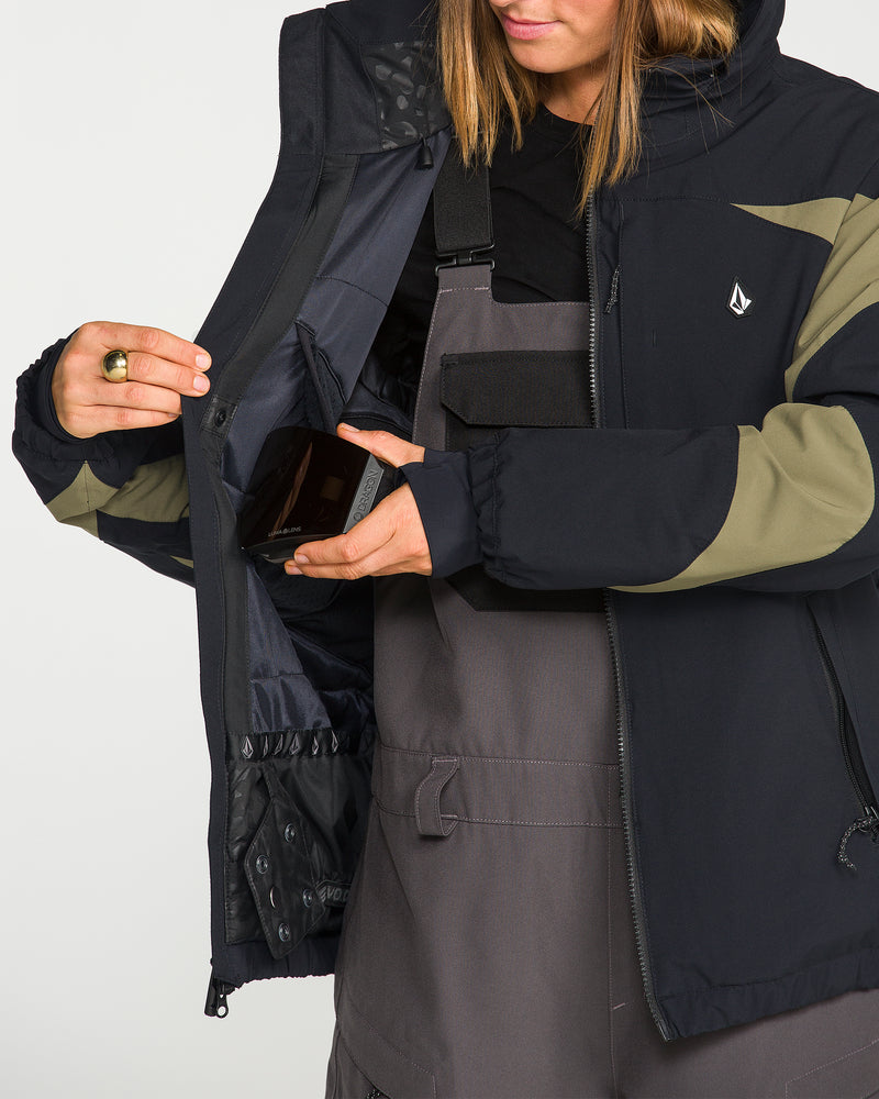 Wearing the Volcom Women’s Shelter 3D Stretch Jacket in black and olive, a person with the jacket unzipped to reveal the inner pocket detail places a smartphone inside, pairing it with gray overalls.