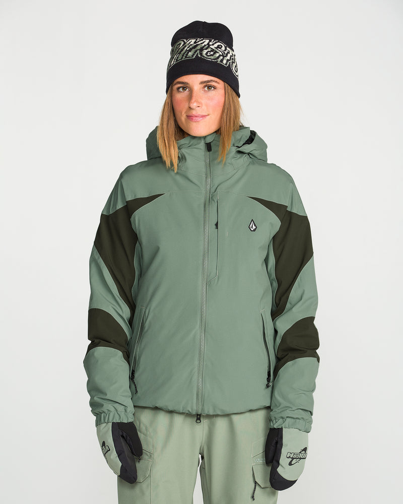 A woman wearing the Volcom Women’s Shelter 3D Stretch Jacket in green and black, featuring waterproof insulation and PFAS-free DWR, paired with matching green pants, black gloves, and a black beanie, stands against a plain white background.
