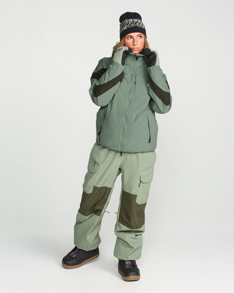 A person is wearing the Volcom Women’s Shelter 3D Stretch Jacket in green with a PFAS-free DWR finish, paired with matching cargo pants featuring dark green patches, black gloves, a black and white beanie, and black boots, all set against a plain white background.