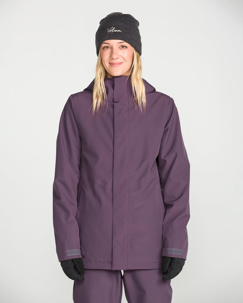 A woman wearing a dark beanie, gloves, and the Volcom Women’s Stoney Shadow Insulated Jacket stands facing the camera with a slight smile against a plain light background.