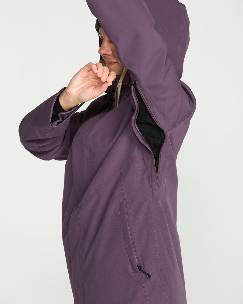 A woman wearing the Volcom Women’s Stoney Shadow Insulated Jacket in purple demonstrates the underarm zipper vent with one arm raised. This insulated ski jacket, made with recycled insulation, is displayed against a plain white background.