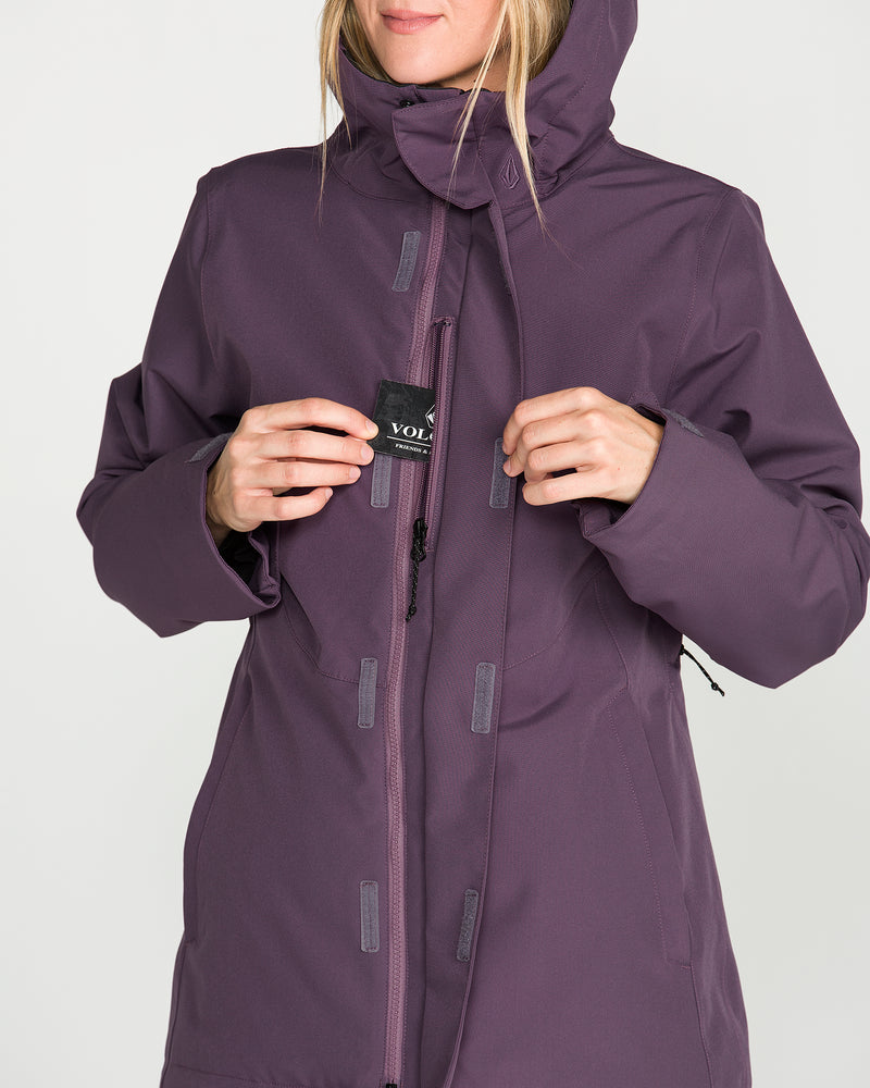 A person wearing a purple Volcom Women’s Stoney Shadow Insulated Jacket is unzipping it and holding a small black tag with “VOL” on it. Their face is partially out of frame, and the background is plain white.